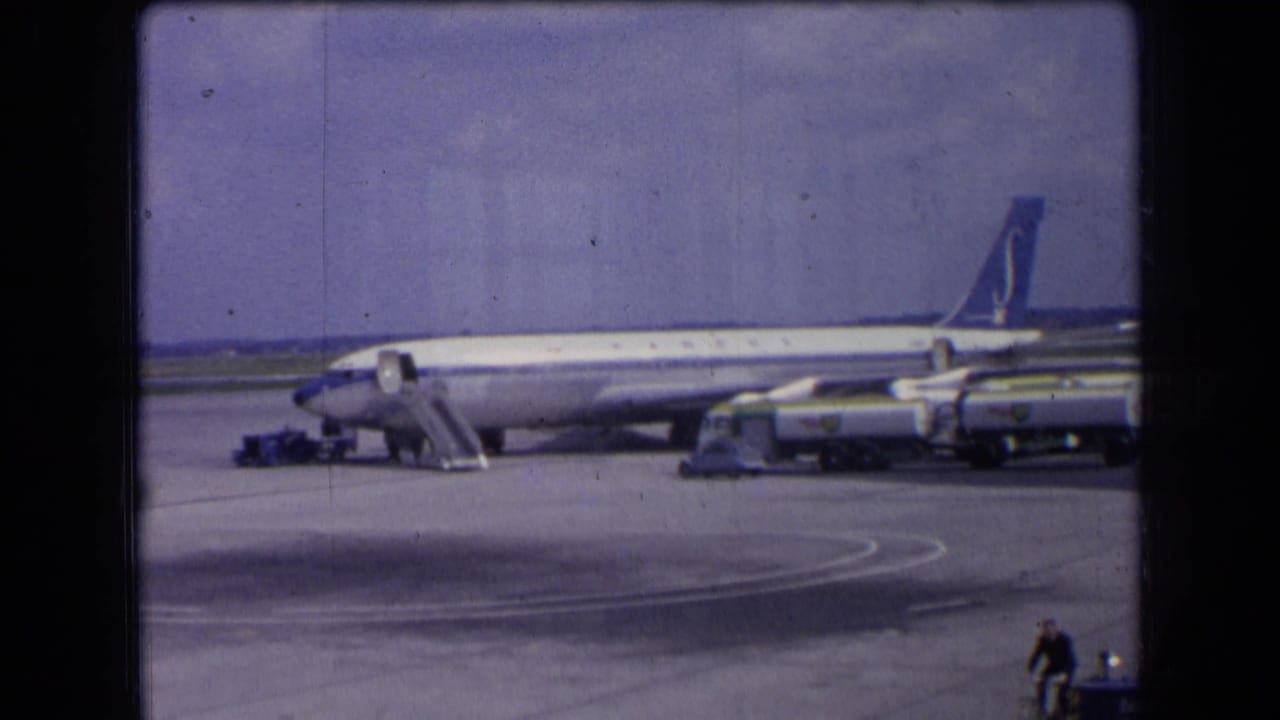 1963 France home-movie frame showing a Sabena passenger jet parked on the tarmac with boarding stairs and ground vehicles under a bright sky.