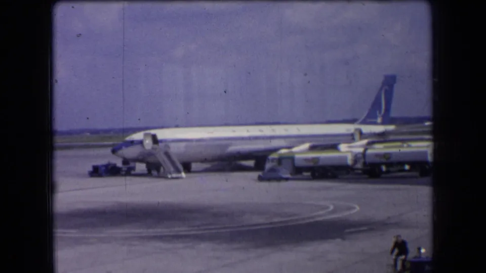 1963 France home-movie frame showing a Sabena passenger jet parked on the tarmac with boarding stairs and ground vehicles under a bright sky.
