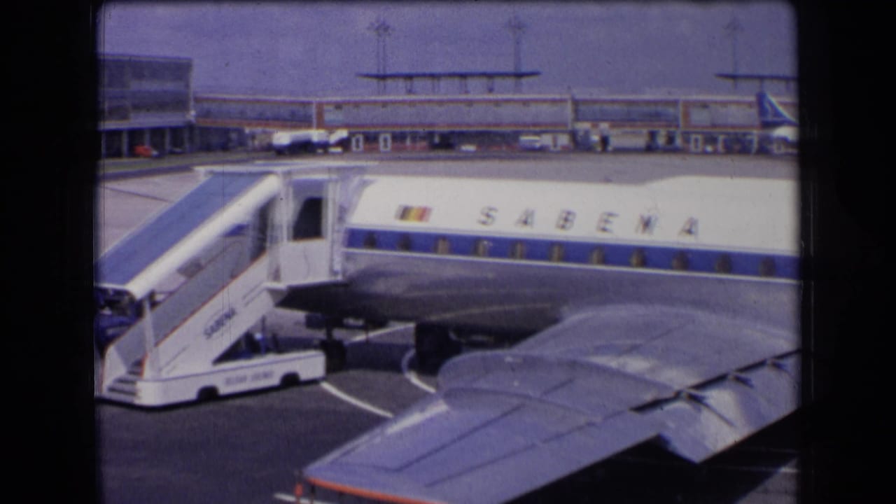 1963 France home-movie frame showing a Sabena aircraft at a gate, boarding stairs attached, with a wing and terminal visible.