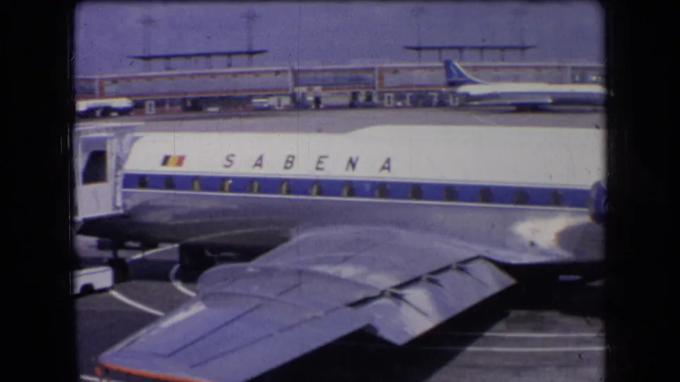 1963 France home-movie frame showing the side of a Sabena aircraft, foreground wing, airport terminal, and distant plane.