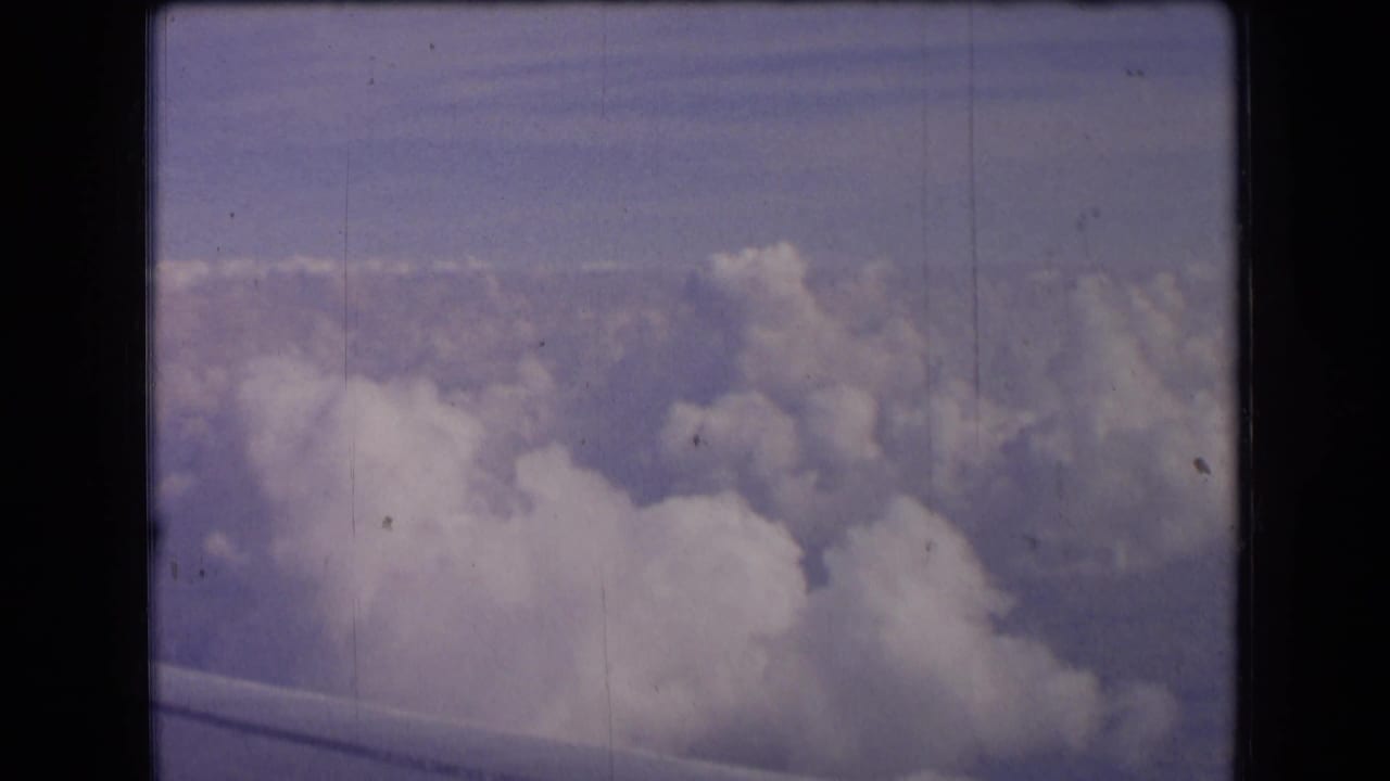 1963 France 8mm frame showing a passenger-window view over clouds, with a partial airplane wing along the bottom edge.