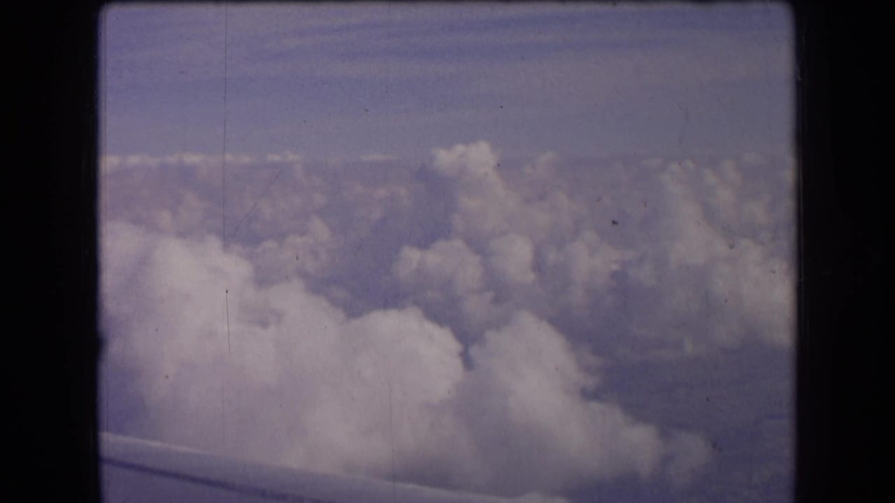 A vintage 8mm passenger view above dense clouds, with the aircraft wing faintly visible at the bottom of the frame.