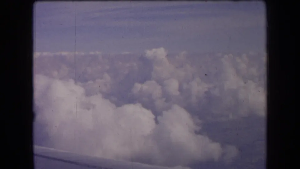A vintage 8mm passenger view above dense clouds, with the aircraft wing faintly visible at the bottom of the frame.