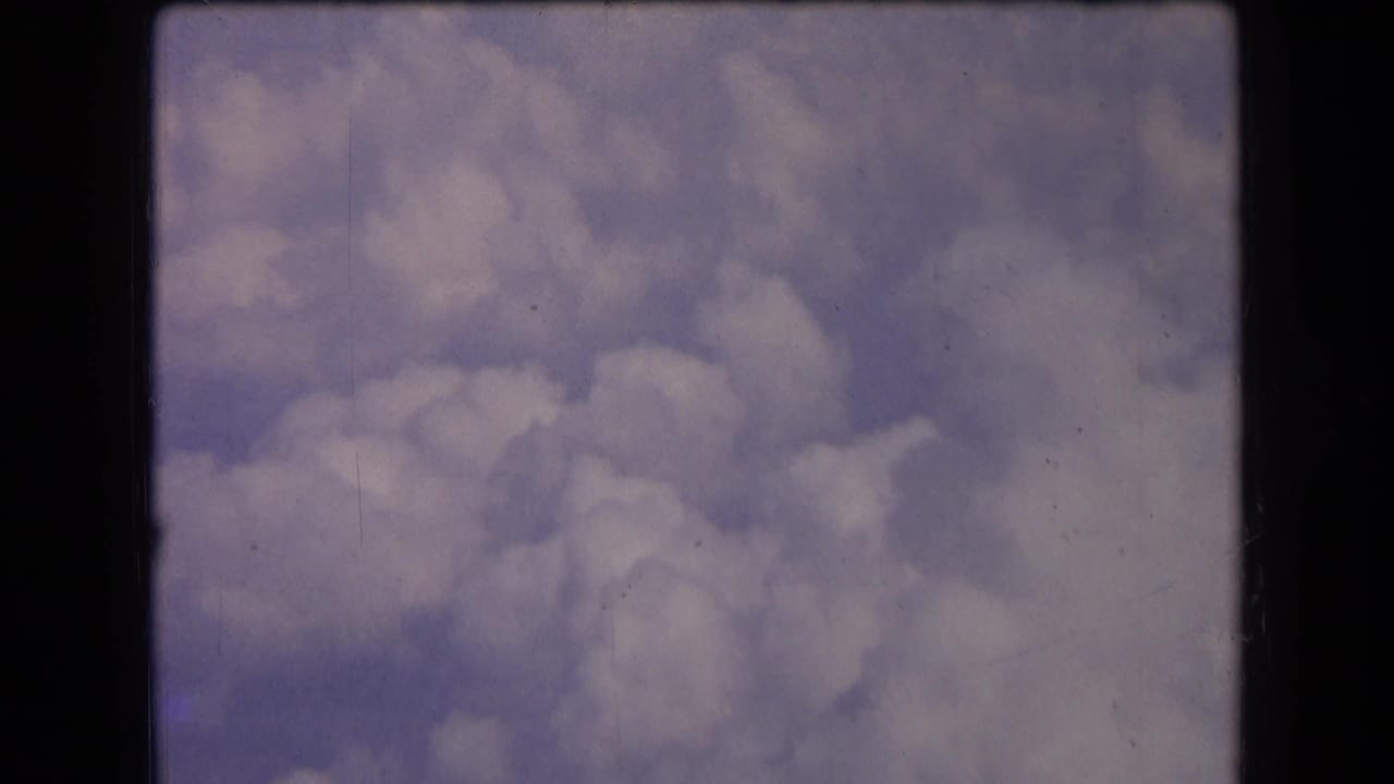 1963 8mm home-movie frame from France showing soft white clouds drifting across a muted blue sky with light grain and dust.