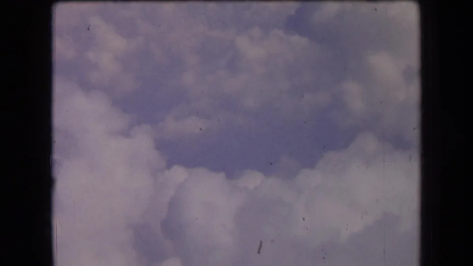 1963 France home-movie frame showing pale blue sky and soft white clouds, marked by grain, dust, scratches, and muted color.
