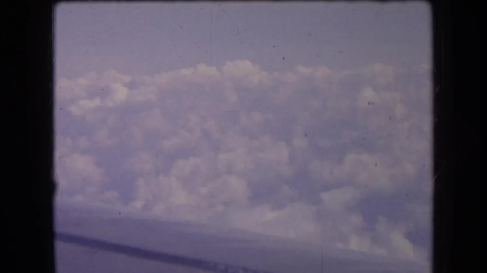1963 France 8mm frame showing pale cloud layers from an airplane window, with muted color and a dark aircraft wing line below.