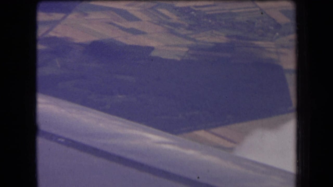 1963 8mm view from an airplane window over rural France, with the wing edge, fields, woodland, and faint clouds below.