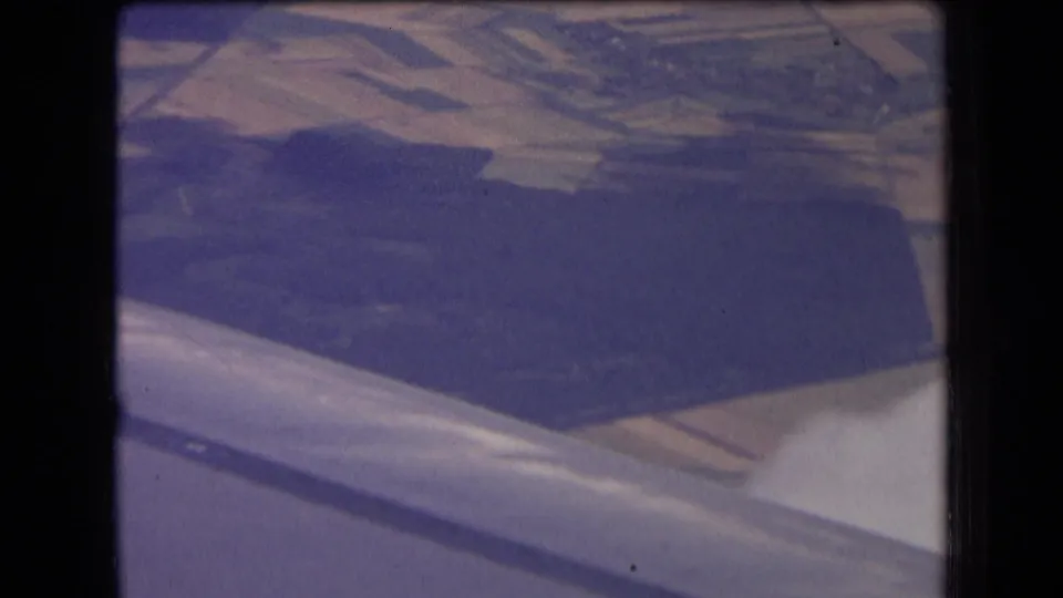 1963 8mm view from an airplane window over rural France, with the wing edge, fields, woodland, and faint clouds below.