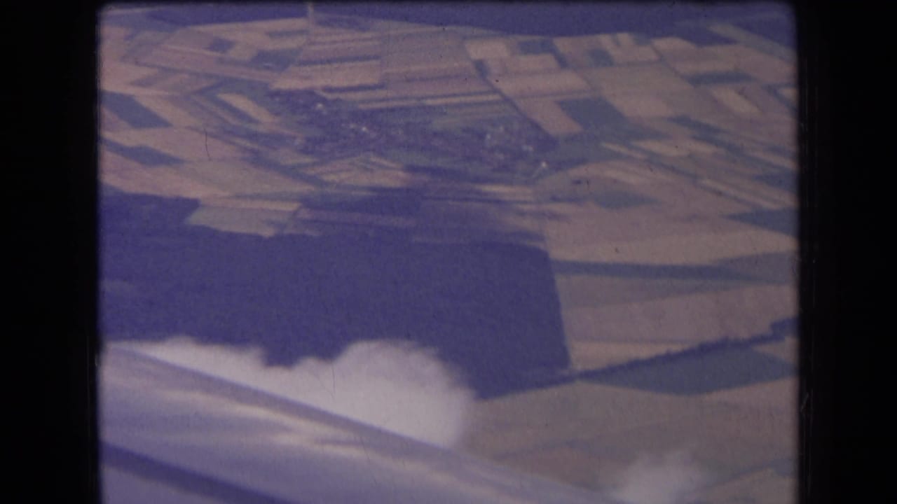Airplane wing crosses the lower frame above fields, woodland, and low clouds.