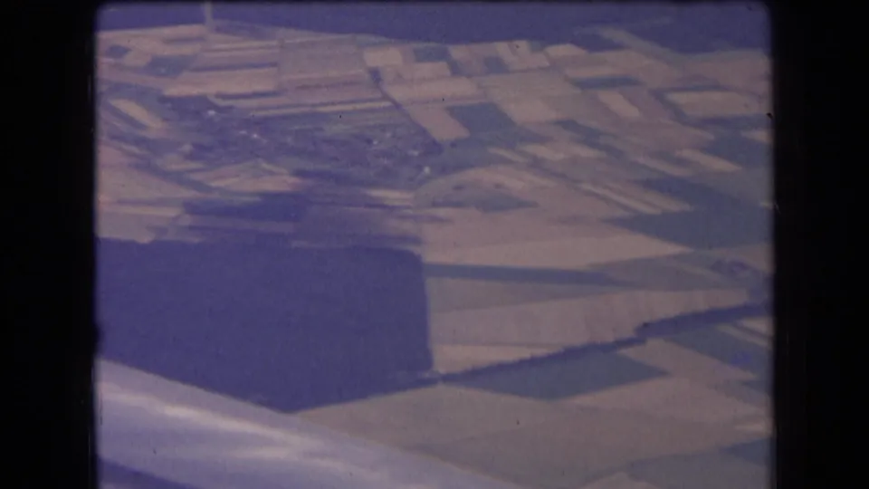 1963 home-movie aerial view over French countryside, showing farmland patterns, a dark wooded block, and the aircraft wing along the bottom.