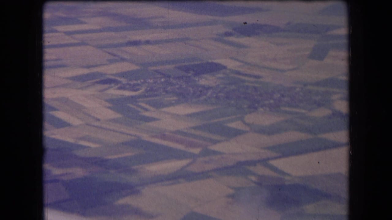 1963 France aerial home-movie frame showing patchwork fields, a distant village cluster, and soft faded 8mm color.