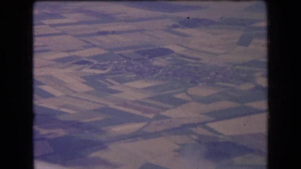 1963 France aerial home-movie frame showing patchwork fields, a distant village cluster, and soft faded 8mm color.