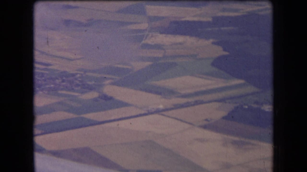 1963 France aerial home-movie frame showing geometric fields, pale plots, and rural roads through soft airplane-window haze.