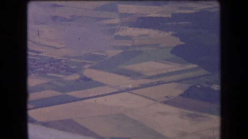 1963 France aerial home-movie frame showing geometric fields, pale plots, and rural roads through soft airplane-window haze.