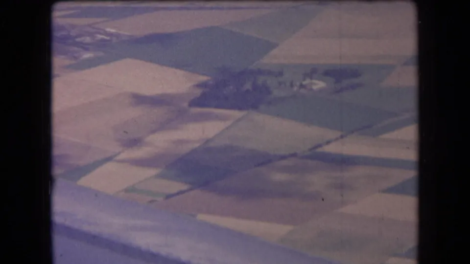 1963 France airplane-window frame with patchwork fields, intersecting roads, and scattered farm structures softened by 8mm grain.