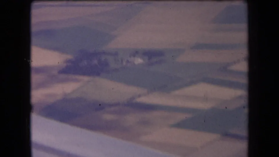 Muted 8mm aerial frame from 1963 France, showing rural fields and a darker central farm area through motion blur and haze.