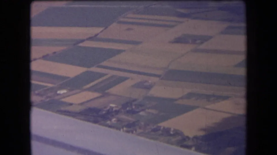 1963 central France aerial footage frame with geometric farmland, isolated farm buildings, tree rows, and the aircraft edge visible below.