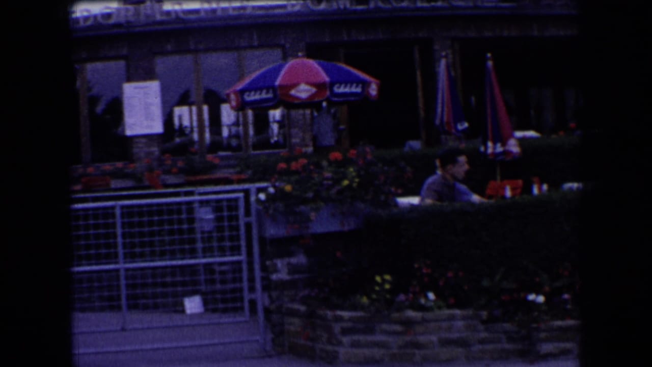 French outdoor café terrace with colored umbrellas, hedges, flowerbeds, and a seated patron near partially visible signage.