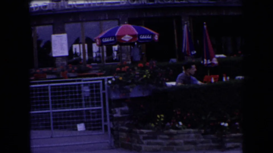 French outdoor café terrace with colored umbrellas, hedges, flowerbeds, and a seated patron near partially visible signage.