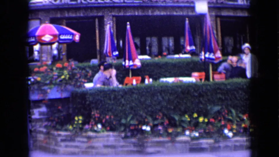 1963 France café terrace scene with patrons seated at tables under umbrellas beside hedges and planted flowers.
