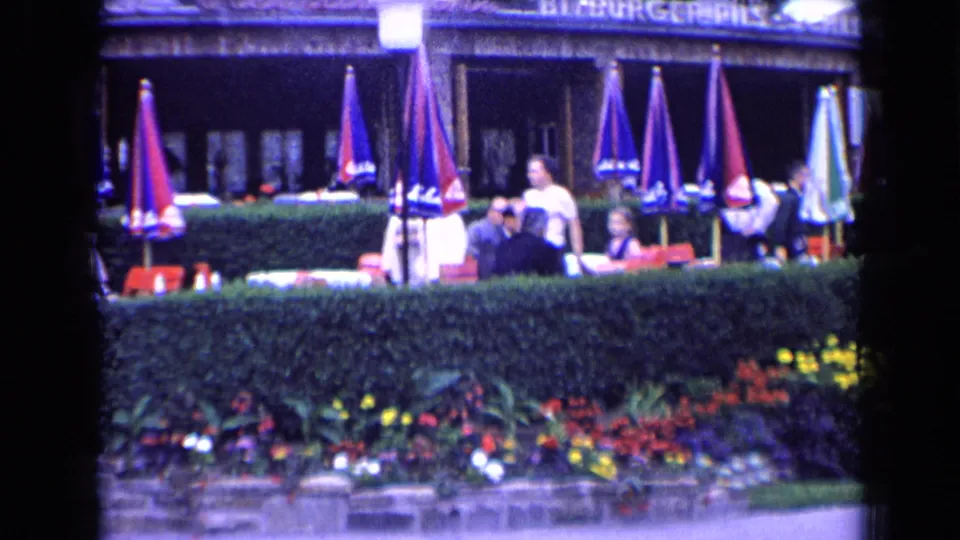 French café terrace with several visible patrons, colorful umbrellas, red chairs, hedges, flowers, and partial storefront signage.