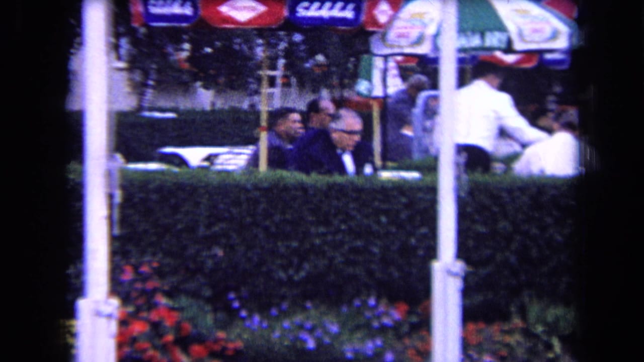 1963 France home-movie frame of adults seated at an outdoor cafe or club terrace behind a hedge, with umbrellas and flowers visible.