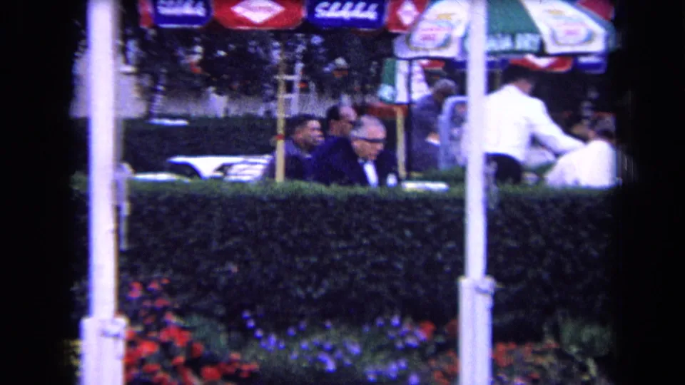 1963 France home-movie frame of adults seated at an outdoor cafe or club terrace behind a hedge, with umbrellas and flowers visible.