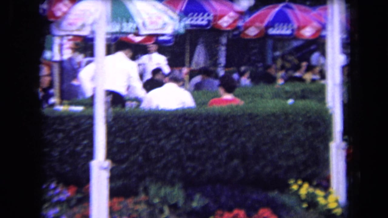 Outdoor terrace scene in France, circa 1963, with patrons at tables behind a hedge and a person in white moving among umbrellas.