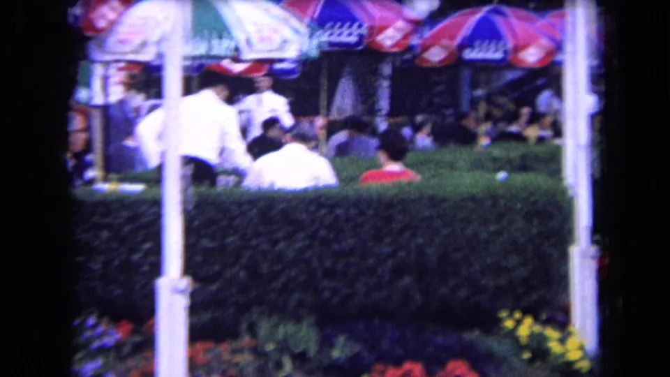Outdoor terrace scene in France, circa 1963, with patrons at tables behind a hedge and a person in white moving among umbrellas.