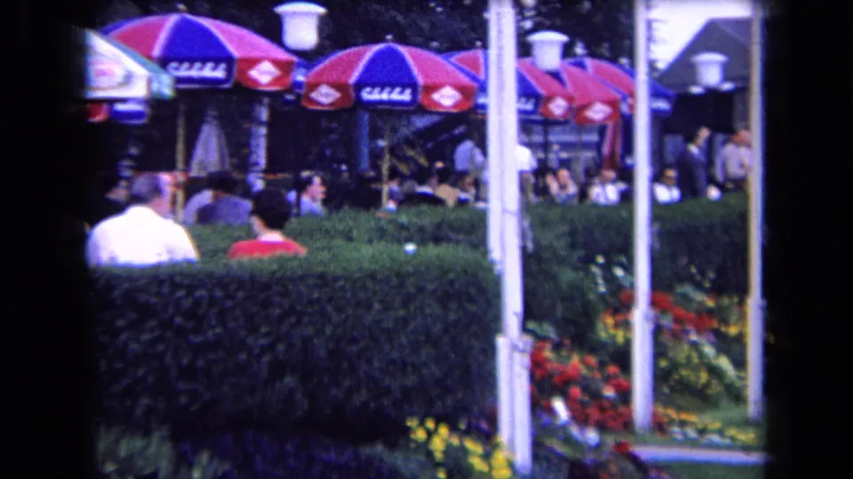 1963 France outdoor cafe or club view with patrons partly obscured by a hedge, flower beds, white posts, and colorful umbrellas.