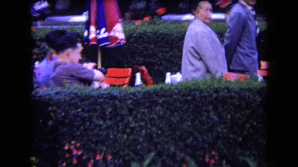 France 1963 frame of adults at an outdoor cafe or club terrace, including a seated woman, standing men, red chairs, umbrellas, and hedges.