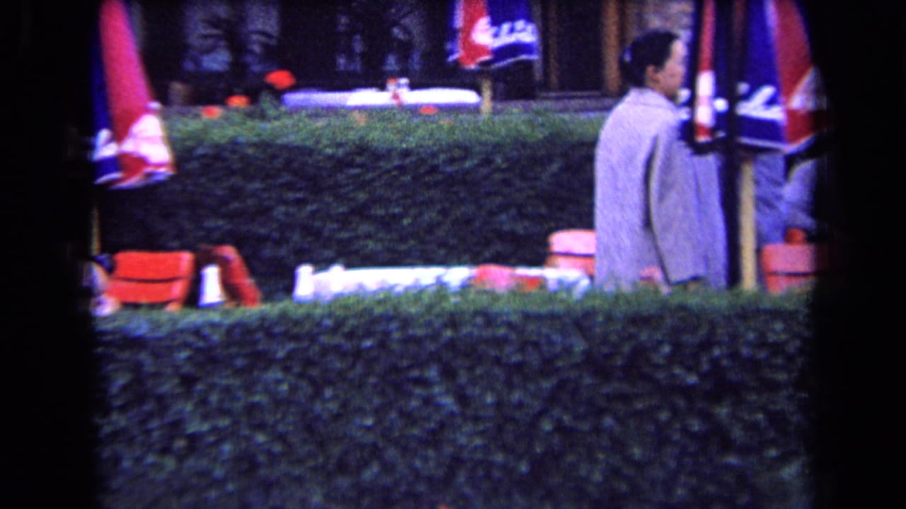 1963 France home-movie frame of a man in a light suit passing an outdoor cafe area with red chairs, umbrellas, and a hedge.