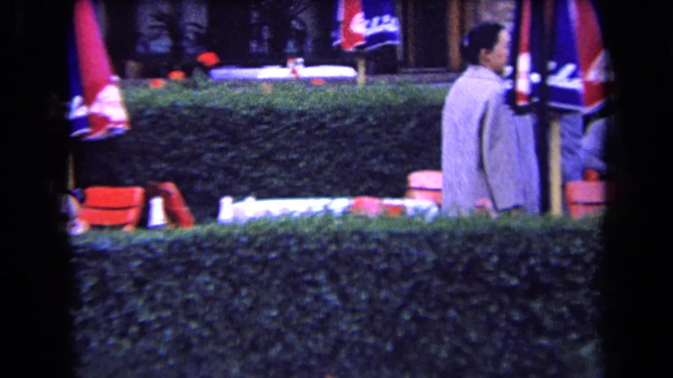 1963 France home-movie frame of a man in a light suit passing an outdoor cafe area with red chairs, umbrellas, and a hedge.
