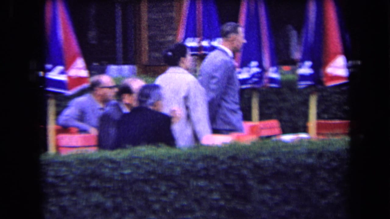 Several adults gather and walk near red cafe chairs, umbrellas, and a hedge.