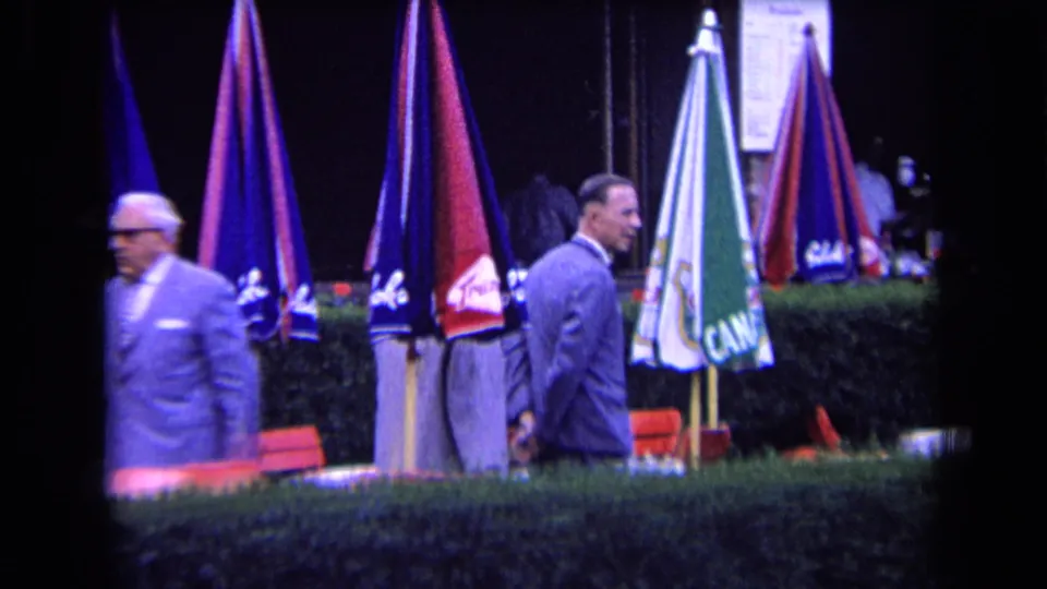 Two men in suits appear beside clustered umbrellas, red chairs, and a hedge in the France 1963 outdoor social scene.