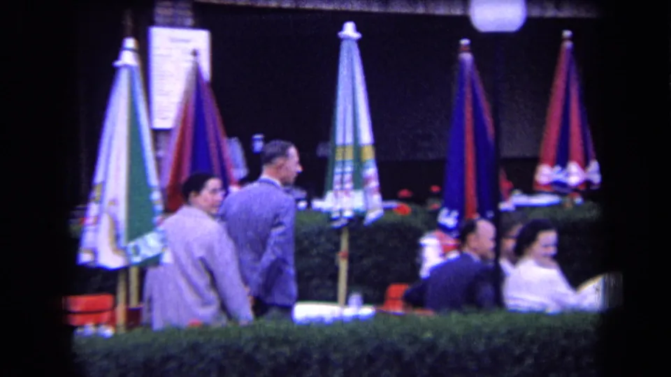 Adults in formal clothing move through a 1963 France outdoor cafe setting with striped umbrellas, red seating, and a hedge foreground.