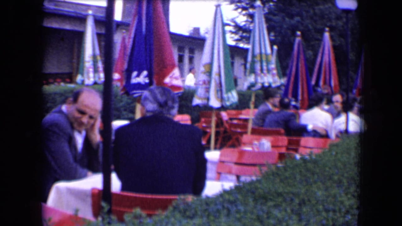 1963 France home-movie frame of people seated at an outdoor café terrace with red chairs and colorful umbrellas behind a hedge.
