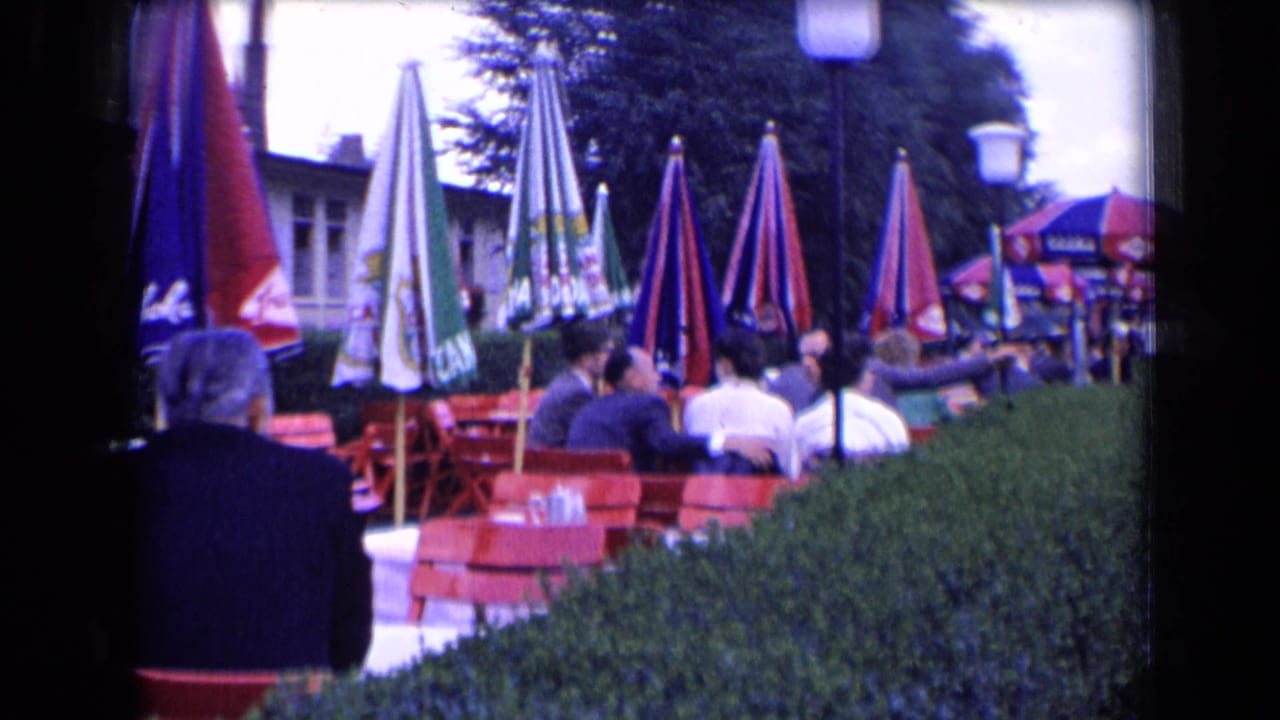 1963 France outdoor café scene with seated patrons, red terrace chairs, umbrellas, hedge, and nearby building façade.