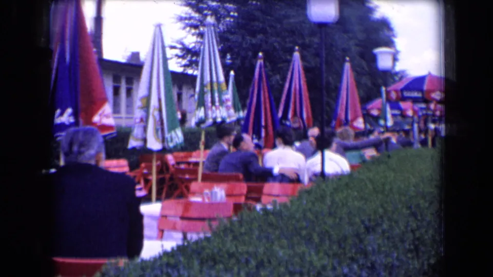 1963 France outdoor café scene with seated patrons, red terrace chairs, umbrellas, hedge, and nearby building façade.