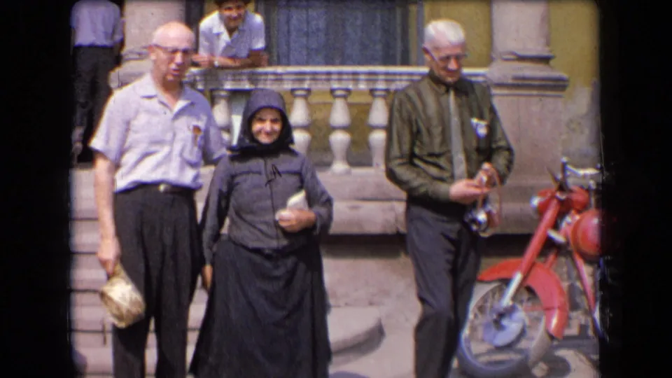 1963 France home-movie frame showing an elderly woman in dark hooded dress escorted by two older men outside a yellow building.