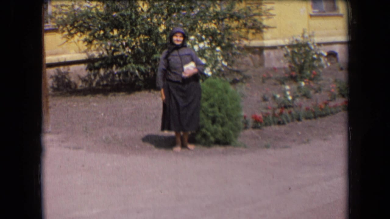 Elderly woman stands alone in a garden area beside shrubs and red flowers.