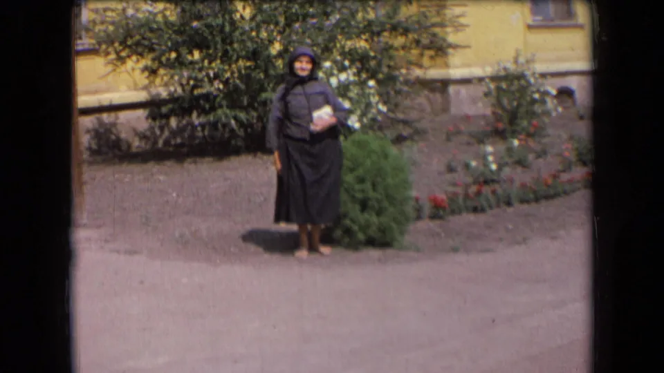1963 France frame of an elderly woman in dark hooded clothing posing alone near bushes and red flowers outside a yellow building.