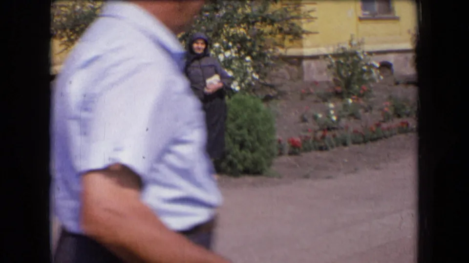 1963 France home-movie frame with a foreground passerby partly blocking the view of the elderly woman standing in the garden.