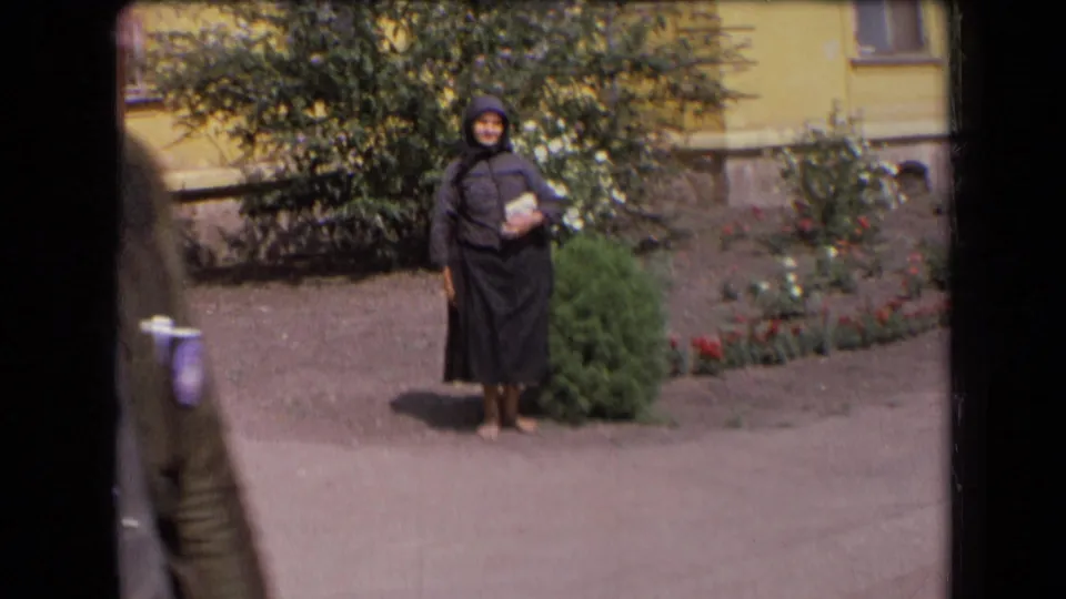 1963 France frame showing the elderly woman in dark hooded clothing near shrubs, with a partial passerby at the left edge.