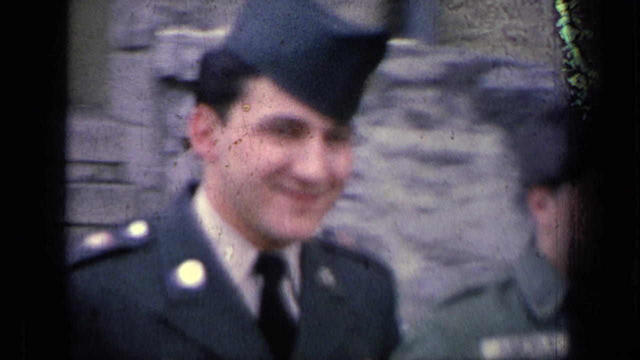 A young U.S. soldier in dark uniform and garrison cap smiles near the camera in soft 8mm footage from the 1963 Germany military-base clip.