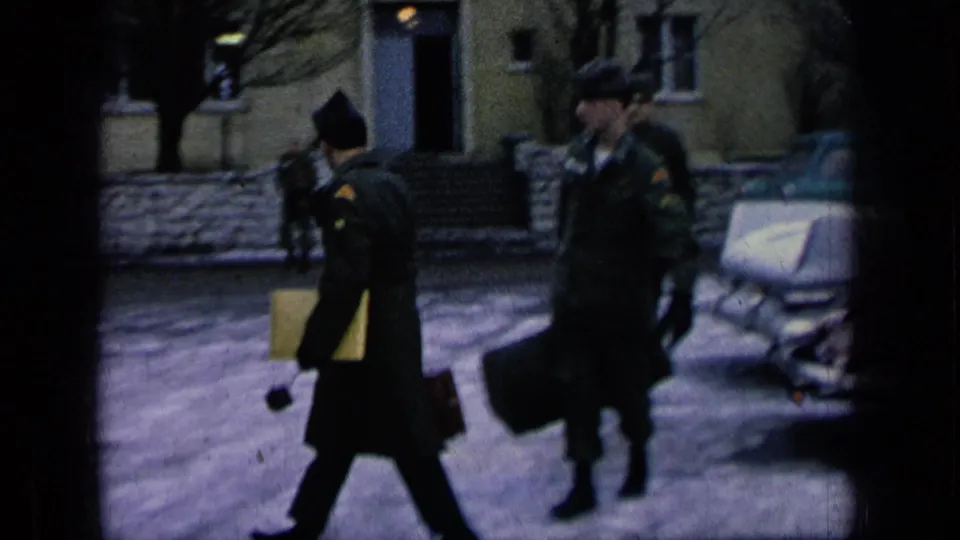 Uniformed soldiers walk across a paved military-base courtyard carrying duffel bags and documents, with cars and a building behind them.