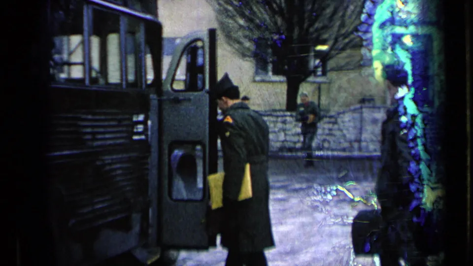 A uniformed soldier approaches an open military bus door in a paved courtyard, with another serviceman and stone wall in the background.