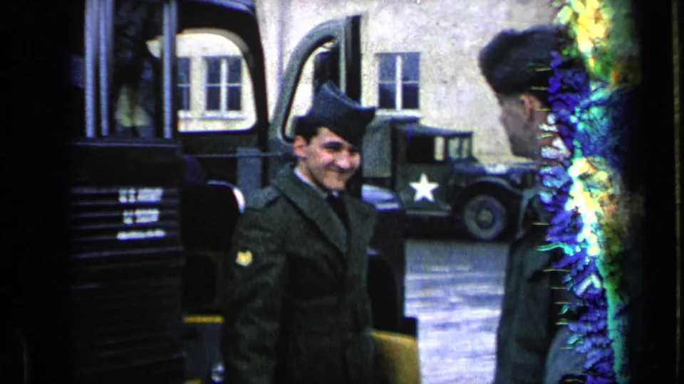 A smiling soldier in uniform stands near the bus, with another serviceman and a U.S. star-marked military vehicle visible in the background.