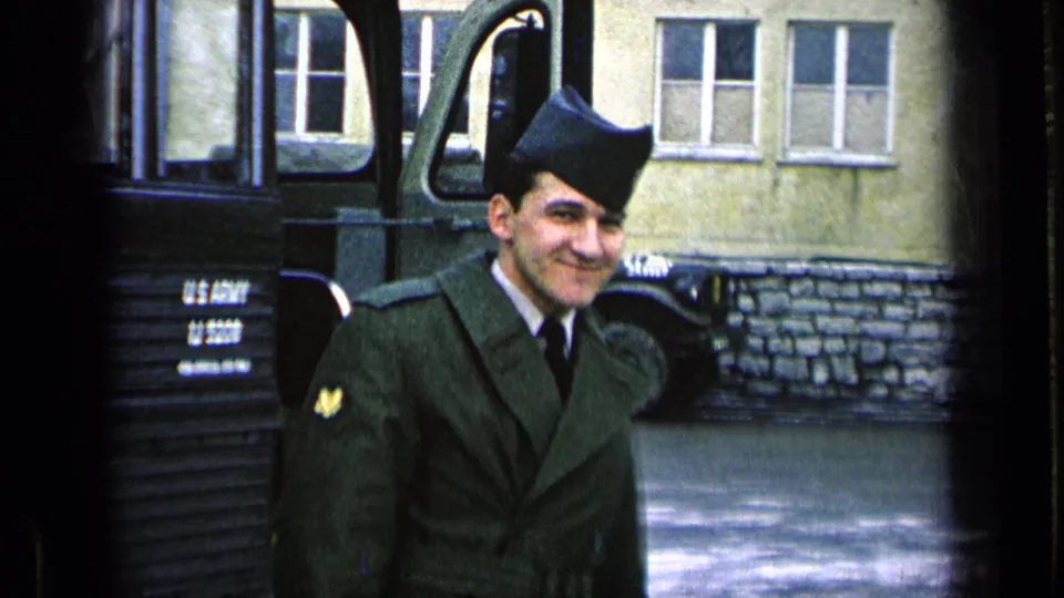 The U.S. Army soldier stands smiling by the military bus, with a base building and star-marked vehicle behind him.