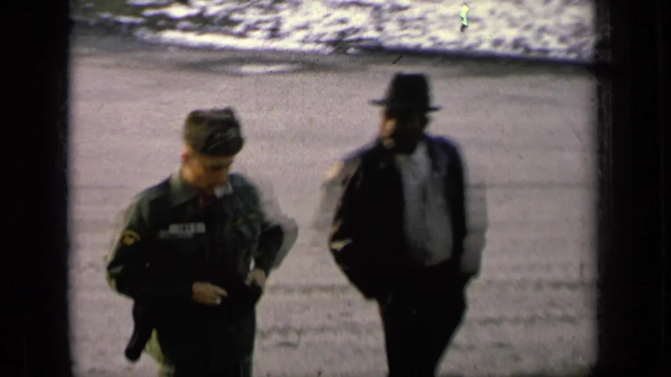 1963 8mm frame of a uniformed soldier walking near a dark-suited man in a fedora at an outdoor military setting.
